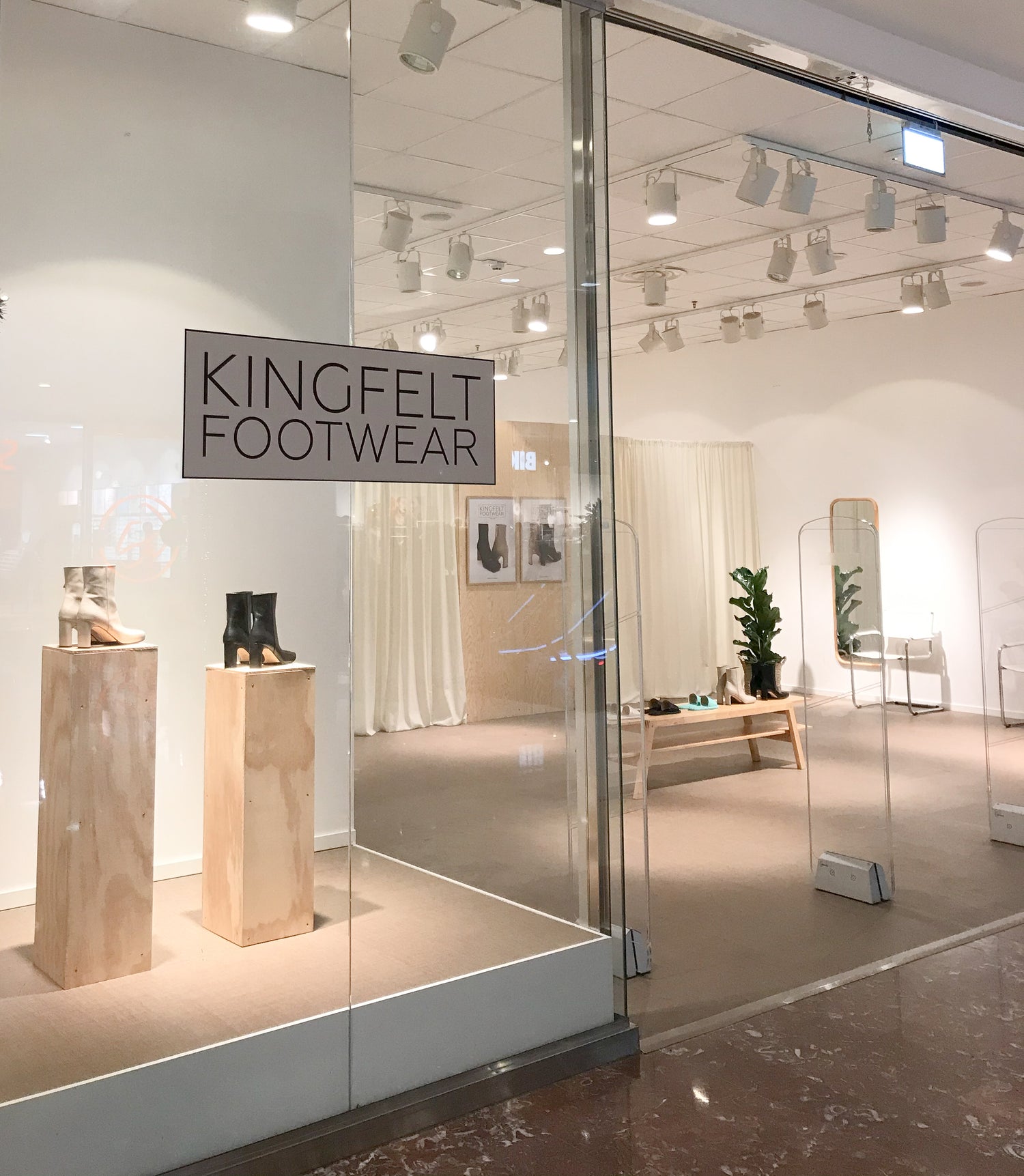 Concept butik KINGFELT FOOTWEAR. Vita cremefärgade sammets gardiner hänger som en avskärmande vägg bredvid en plywood vägg med afficher med skinnstövletter på. Två pidestaler i plywood i skyltfönstret med square toe boots stövletterna på. 